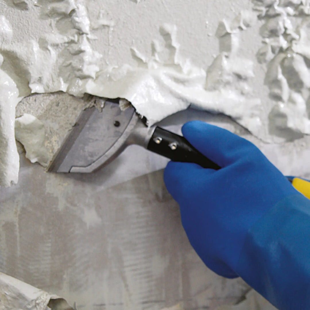 Hand wearing a blue glove applying white putty to a wall with a putty knife.