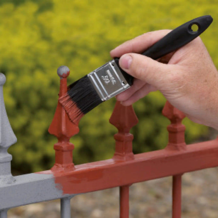 Hand holding a paintbrush applying red paint to a metal fence with a blurred green background