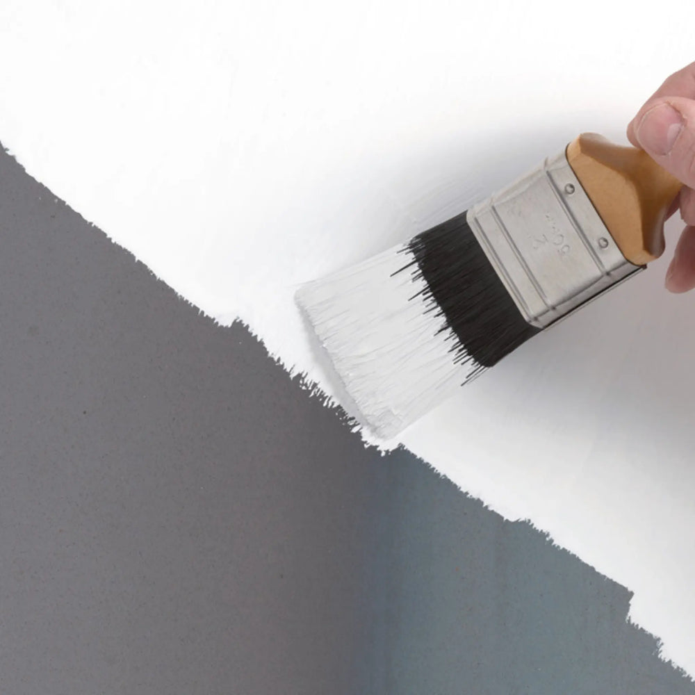 Person painting a wall with a brush, transitioning from white to gray.