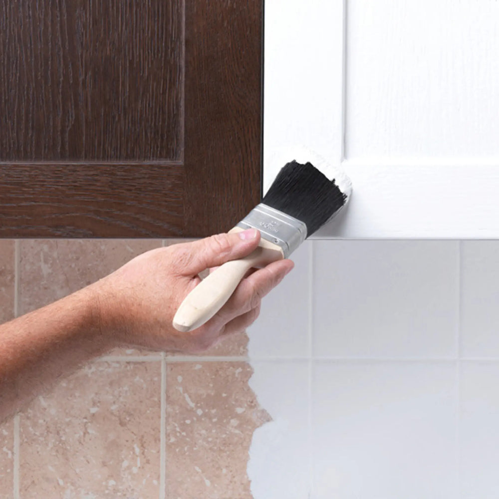 Hand using a paintbrush to apply white paint to a corner of a room with tiled walls and a wooden door.