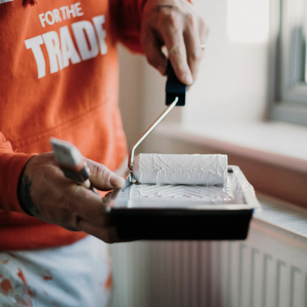 Person holding a paint roller tray with a paint can and roller, wearing an orange shirt with text.