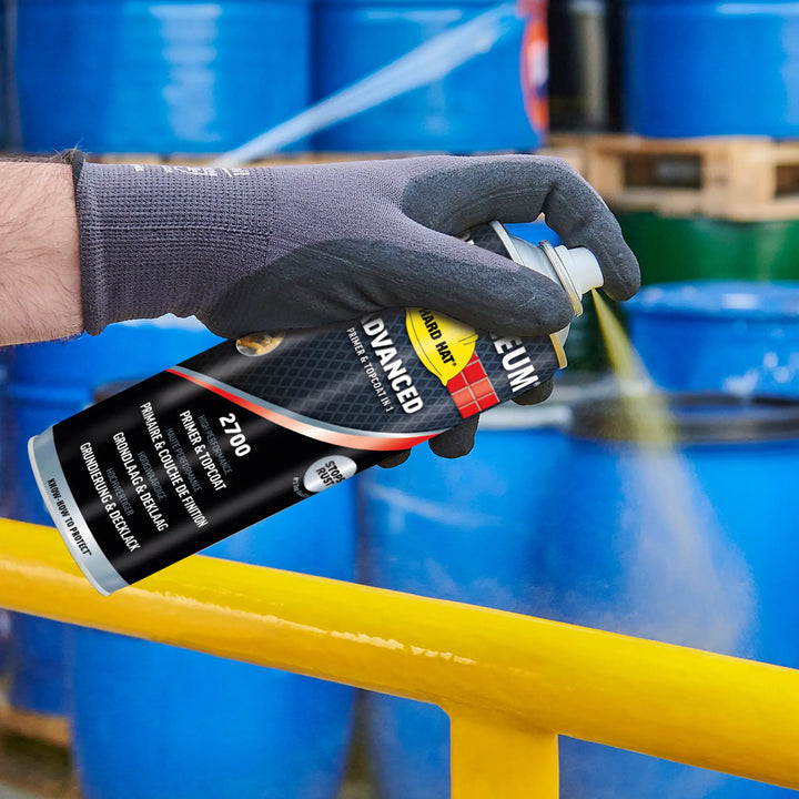 Hand wearing a black glove holding a can of spray paint with blue barrels in the background